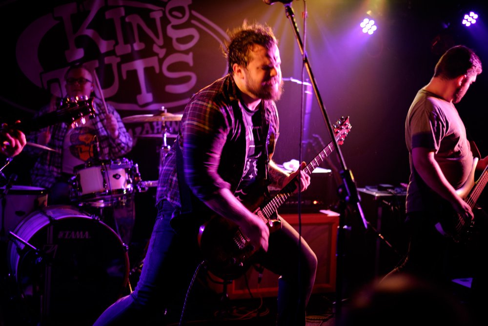 IKARI's lead singer Kris rocking out on stage. To either side you can see the drummer, Chris, and the bassist, Kris. Behind them all is a sign saying 'King Tut's'