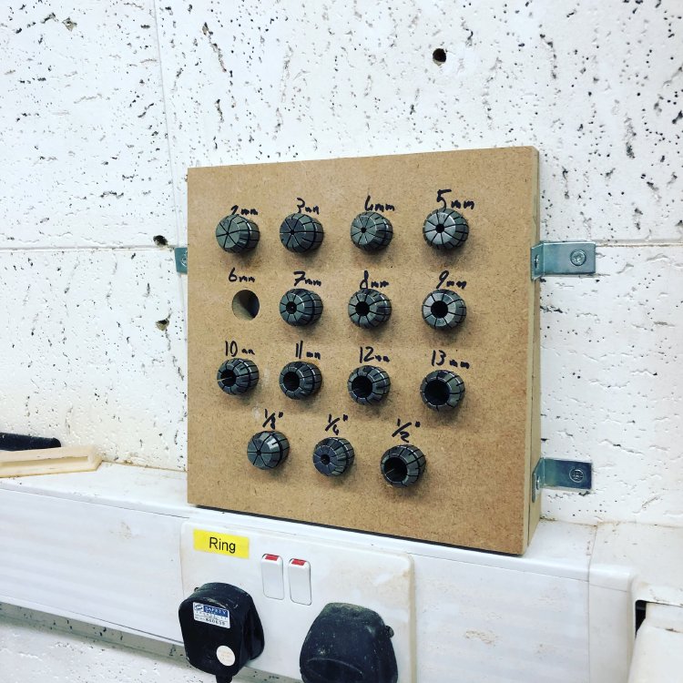 A photo of a workshop organisation board made from MDF mounted on a white wall, displaying a collection of precision collets arranged in a grid pattern and labeled with their sizes (2mm through 13mm, plus fractional sizes), with metal clips securing the board to the wall.