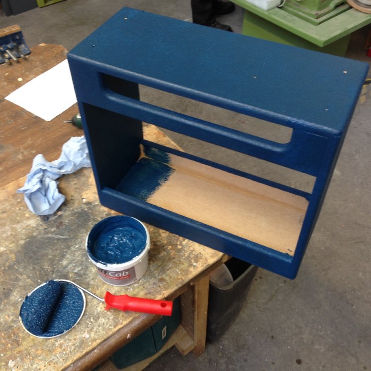 A empty amp cabinet sits on the workbench being painted with a blue substance. You can see a small roller and tub of paint beside it.