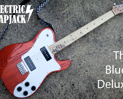 An electric guitar sits on a concrete background. It has an orange body with a white pickguard and black P90 pickups and controls. The neck is pale like maple, with a black figuring in the lower half.