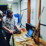 A photo of me stood by a table with guitars and a laptop on, explaining to someone how guitars are designed and built.