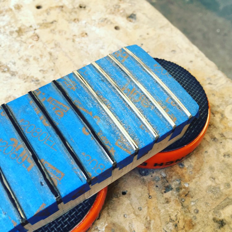 A guitar neck sits on the workbench, with the fretboard covered in masking tape to leave just the ferts showing. Half the frets have black sharpie on them, waiting to be shaped, the rest are shiny where they've been worked on.