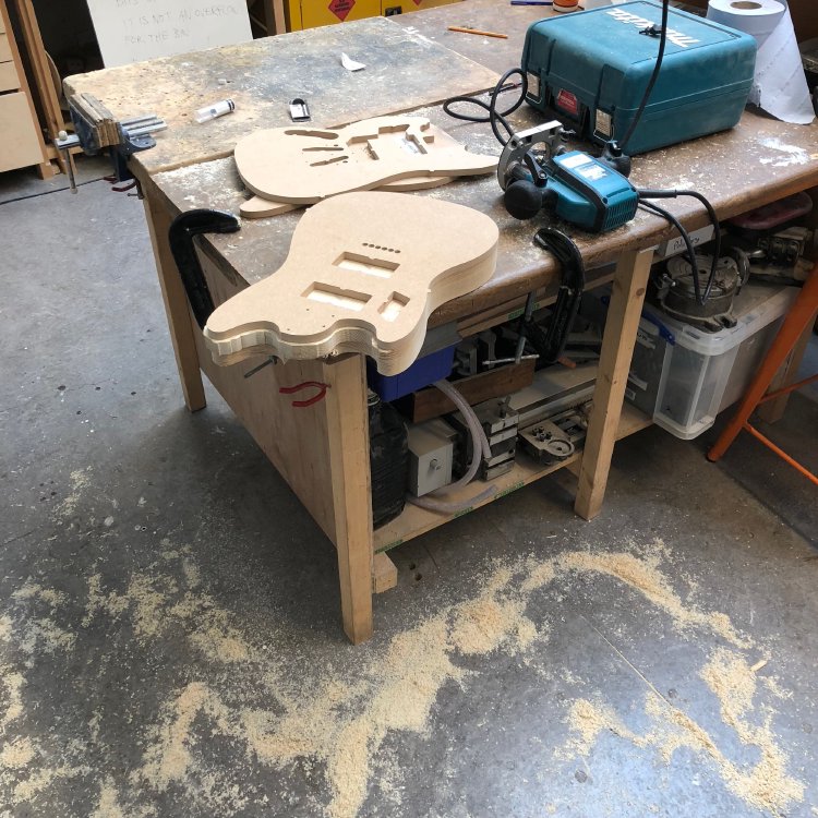 A guitar body being shaped using a hand router sites part cut on the workbench. The floor is covered with wood dust.