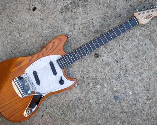 A photo of a solid-body offset style electric guitar laid on a concrete background. The body is natural wood finished, stained orange, it has a white pearlescent pick-guard, into which are set two single coil pickups and a rotary pickup selector switch. It has a tremolo unit in the bridge. The neck is maple with a wenge fretboard.