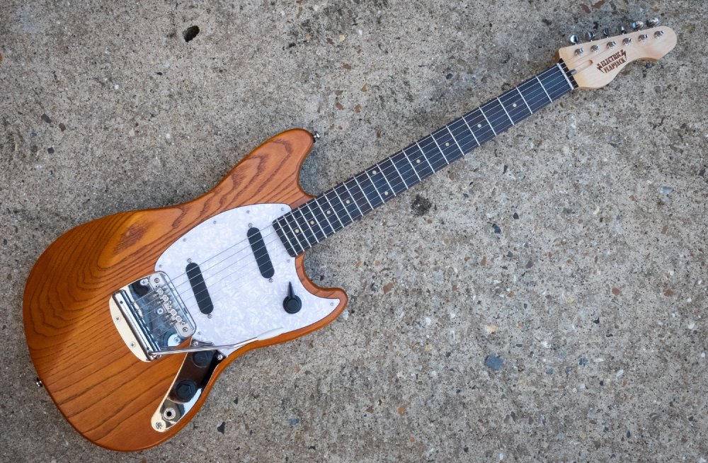 A photo of a solid-body offset style electric guitar laid on a concrete background. The body is natural wood finished, stained orange, it has a white pearlescent pick-guard, into which are set two single coil pickups and a rotary pickup selector switch. It has a tremolo unit in the bridge. The neck is maple with a wenge fretboard.