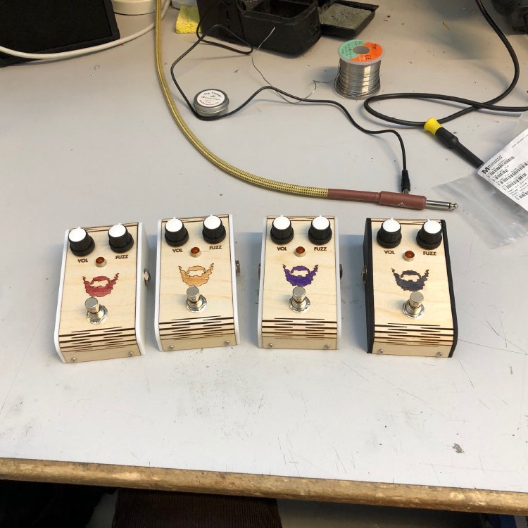 A row of four guitar effects pedals sits on the workbench. Each one has a laser-etched wooden face with a beard logo, each pedal having the beard stained a different colour.