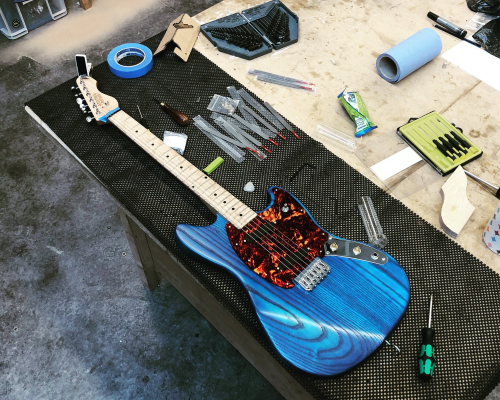 A striking blue electric guitar with a red tort pickguard and maple neck sits on the workbench as it undergoes final tweaking.