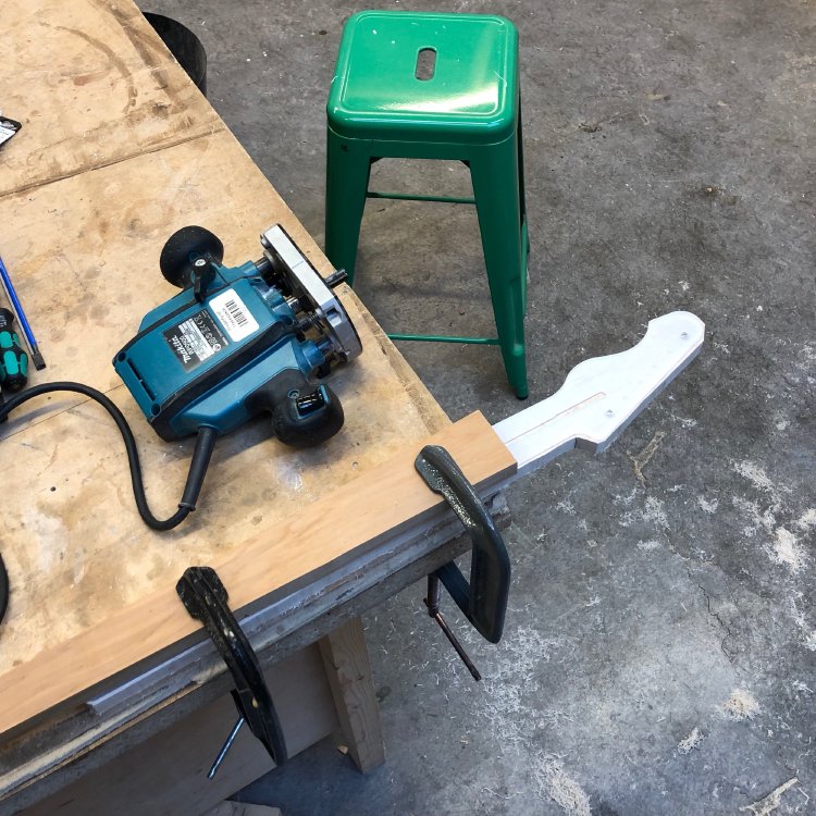 A crudely shaped guitar neck, with template still attached, is clamped to the workbench. Beside it is a plunge-router.