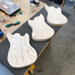 Three early stage guitar bodies, all just cut out as profiles from blocks of wood, sit on the workbench in a row.