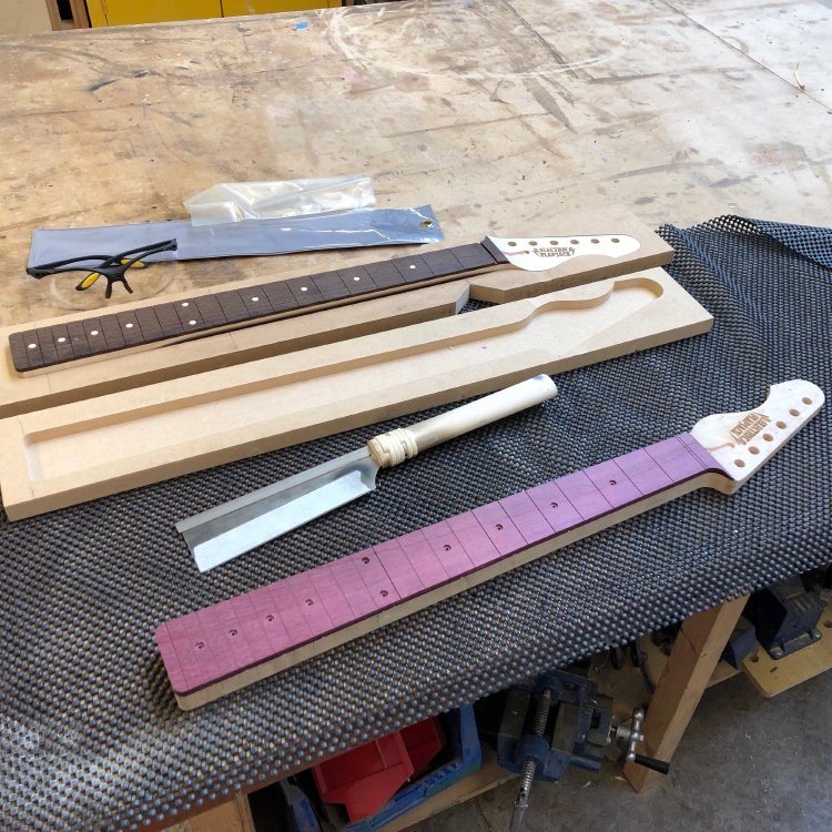 A view of two in progress guitar necks on the workbench. Both one is maple with a wenge fretboard with maple inlays. It has the fret slots cut, the fretboard is radiused, and the back of the neck is carved. The other neck is maple with a purpleheart wood fretboard. It has the fret slots cut and insets for the inlays drilled, but has not been either radiused or carved on the rear.