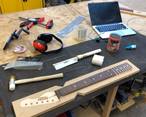 A messy workbench shows a guitar neck in late build process, with about a third of the frets inserted. Beside the neck sits a hammer with plastic ends, a fret-slot saw, a laptop, some fret-wire, and misc other tools.