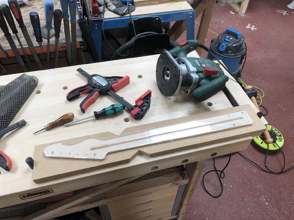 A work in progress guitar neck sits in a jig with a template on it upon a workbench. Beside it sits a plunge-router, some clamps, and a screwdriver.