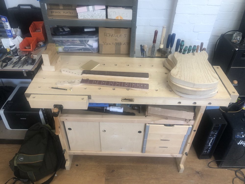 My sjöbergs workbench sits in a brightly lit workshop, and upon it are two un-finished guitar necks and three un-finished guitar bodies. Besides them sits a notebook and a pencil.