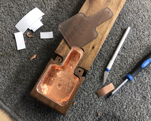 The rear body of the guitar on a carpet mat on a workbench, showing the copper covered inside of the electronics cavity. To the side you can see a scalpel and bits of tape, and on the body to one side you can see the translucent orange acrylic cover for the cavity that will let the shiny copper inside be visible once the cover is on.