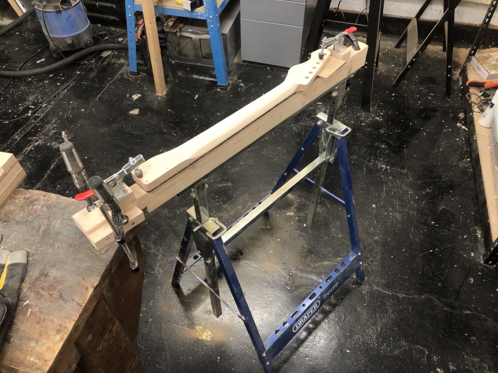 What looks roughly like the neck of a guitar sits on a trellis frame next to a workbench, ready to be worked on.