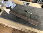 A work in progress guitar neck, shaped and fretted, sat on a workbench. Beside the neck is a black sharpie pen, and closer inspection reveals all the frets have been drawn on with the sharpie ready for fret shaping work.