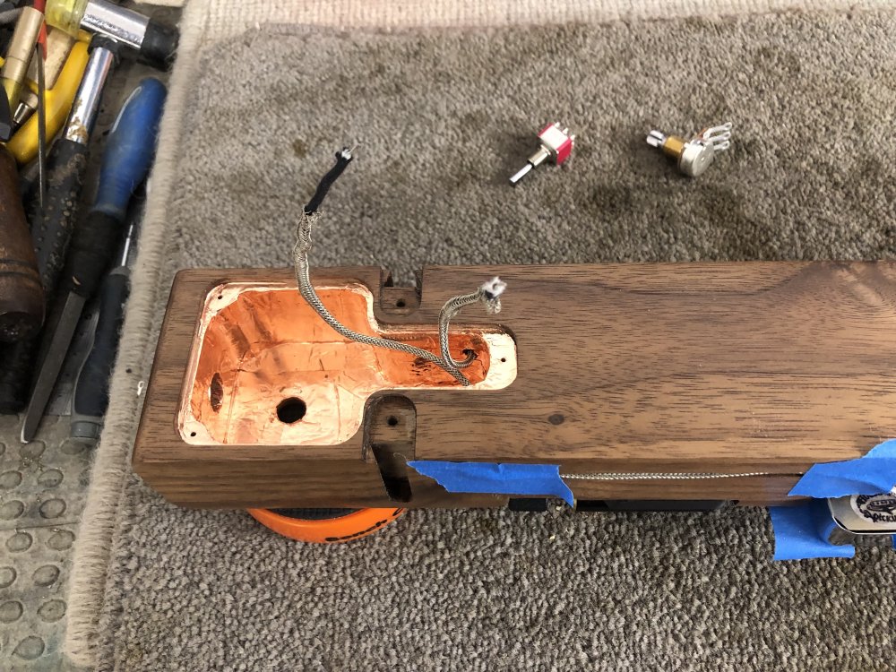 The tail of the guitar with its back up, so you can see the copper-tape lined cavity for the electronics. Two pickup cables are poking out at odd angles, and you can see a switch and a potentiometer in the background.