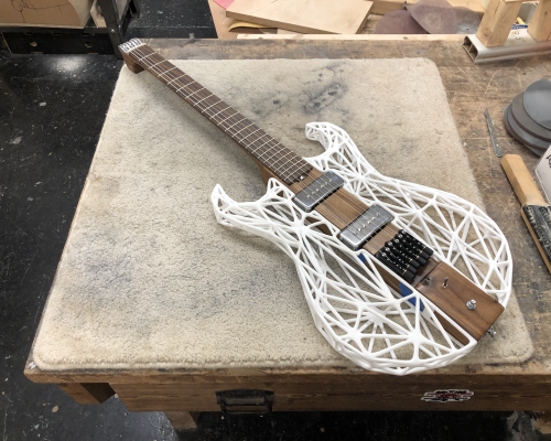 An unusual looking guitar sat on a workbench. The guitar consists of a wooden central core that consists of the neck extends that profile through the body, and then the sides of the body are made from a 3D printed lattice. The guitar has no headstock, instead having tuners at the bridge.