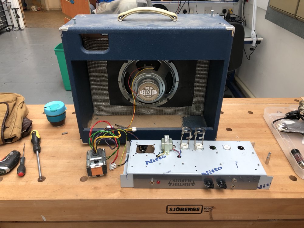 A workbench upon which sits the amplifier, facing backwards, with the metal chassis with the electricals in sat beside it, and the original power transformer removed sat next to these. The top of the amp is very dusty from having been kept in a maker-space for 18 months.