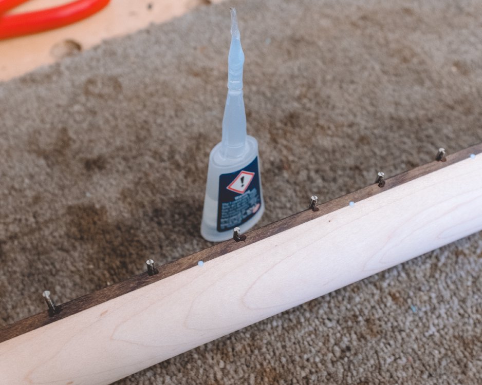 The guitar neck sits on its side, next to a bottle of super glue that has a wicking narrow spout on it. The edges of the frets stick out beyond the side of the neck, and you can see a damp patch of glue on the neck where the fret juts out.