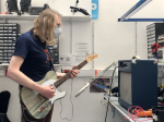 I'm stood next to the workbench with a blue guitar amp on it, and I have a guitar plugged into the amp for testing purposes. I'm staring at the amp as I play.