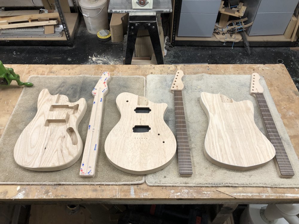 Three work in progress guitars sit on a workbench, each a body with a neck beside it. All are raw wood and unfinished. The left body has pickup cavities and a neck pocket cut, and the neck is just roughly shaped with a template on top. The middle one is the most complete, with the neck fretted and all the body holes cut. The right body has only a neck pocket but no other holes init, but the neck is fretted and otherwise complete.