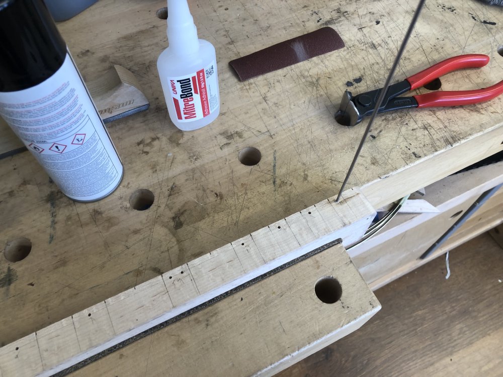 A view of a guitar neck in a vice showing the player facing side up. Holes have been drilled at the 3rd, 5th, 7th, 9th, 12th, 15th, 17th, 19th, and 21st frets ready for dowel to be inserted, and the rod of metal dowel is currently stuck into the 21st hole.