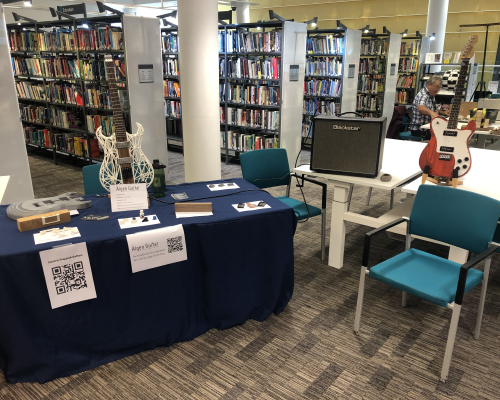 A photo of a set of tables made up to display my Älgen hybrid built guitar (part wood, part 3D-printed), with examples of prototype parts, wood off cuts, and an amplified ready to let people try it out.