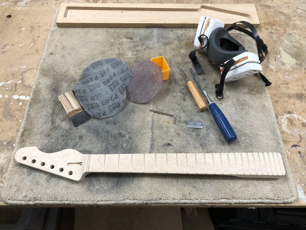 An overview of the neck on the workbench, surrounded by sanding disks, chisels, my respirator, a razor blade, and an allen-key.