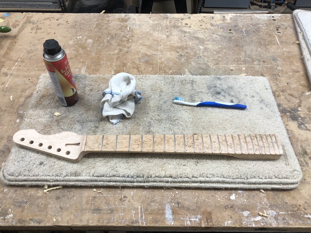 A guitar neck sits on the workbench on a carpet tile for protection, and next to it is a bottle of 'liquid gold' cleaner, a white cloth, and a tooth-brush.