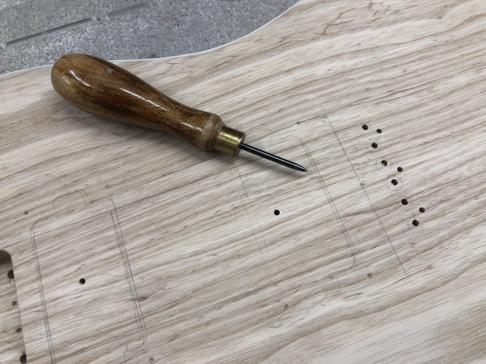 A close up of the guitar body, where you can see the pickup cavity positions and the mounting screw holes around them have been marked out in pencil. Atop the body is a bradawl, a small tool with a wooden handle and then a metal shaft that comes to a point.