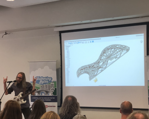 A photo of me talking in front of an audience. I'm stood wearing a guitar on a strap in front of a slide showing a CAD model of the same guitar, and I'm doing the devil's horns gesture with my hands for some reason.