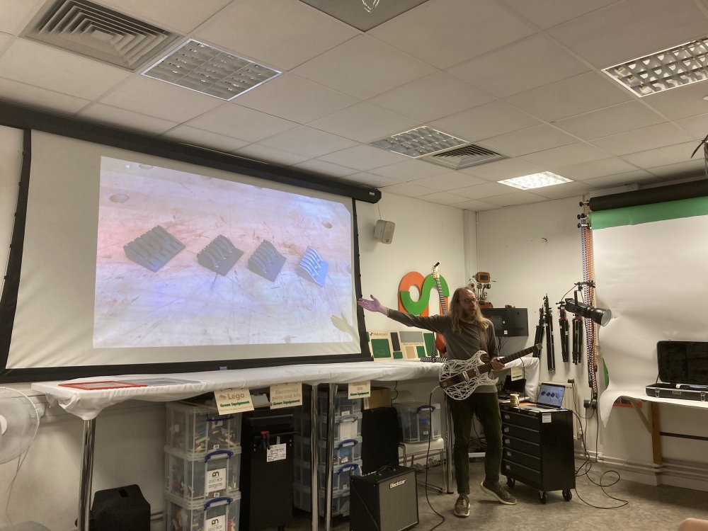 A photo of me in the Makespace classroom, holding my Älgen wood and 3D-printed guitar in front of a projected slide showing the evolution of the 3D-printed metal headstock part, showing the three different plastic prototypes and the final metal version.