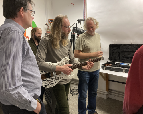 A photo of me holding my Älgen wood and 3D-printed guitar in a cluster of onlookers. I look like I've escaped from a 70s edition of the open university programs.