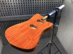 An orange-stained guitar body sits mounted on a jig in a spray booth.