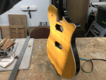 A photo of a guitar body being held over a workbench, caught in the sunlight coming through an unseen window. The face of the body is yellow stained birds-eye maple, and the top 5mm of the side is natural unfinished birds-eye maple, and then the back of the guitar is painted gloss black.