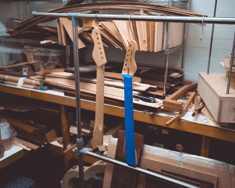 A photo of two guitar necks hung up on rack in room otherwise filled with wood on shelves. Both necks are made from maple: one is hung so you can see the back, showing the birds-eye figuring, and the other is hung so you can see the fretboard is covered in masking tape.