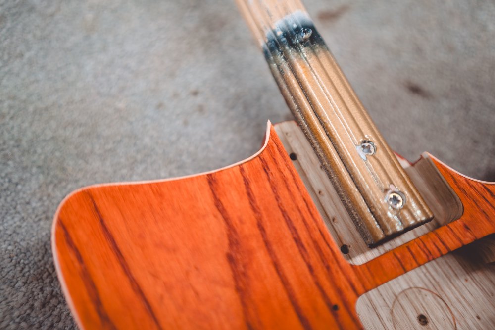 A close-up photo of the neck pocket on the orange-stained body, where  you can see the tight corners of the body into the neck, and in there the white plastic binding that goes around the top edge is pulling away from the body.