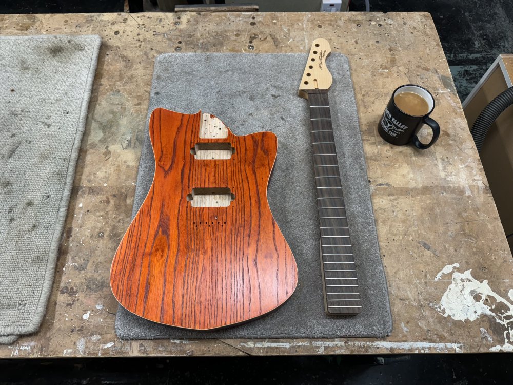 A photo of a workbench, atop of which is the orange stained body with its in-place binding, next to which sits the associated maple and rosewood neck, and next to which is a mug of tea.