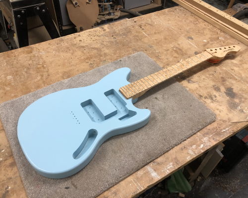 On the workbench sits a work-in-progress guitar, with just the painted-body and neck in position. The body is in the style of a Fender Mustang offset guitar and is painted a pale blue, and the neck is made from birds-eye maple and finished in oil.