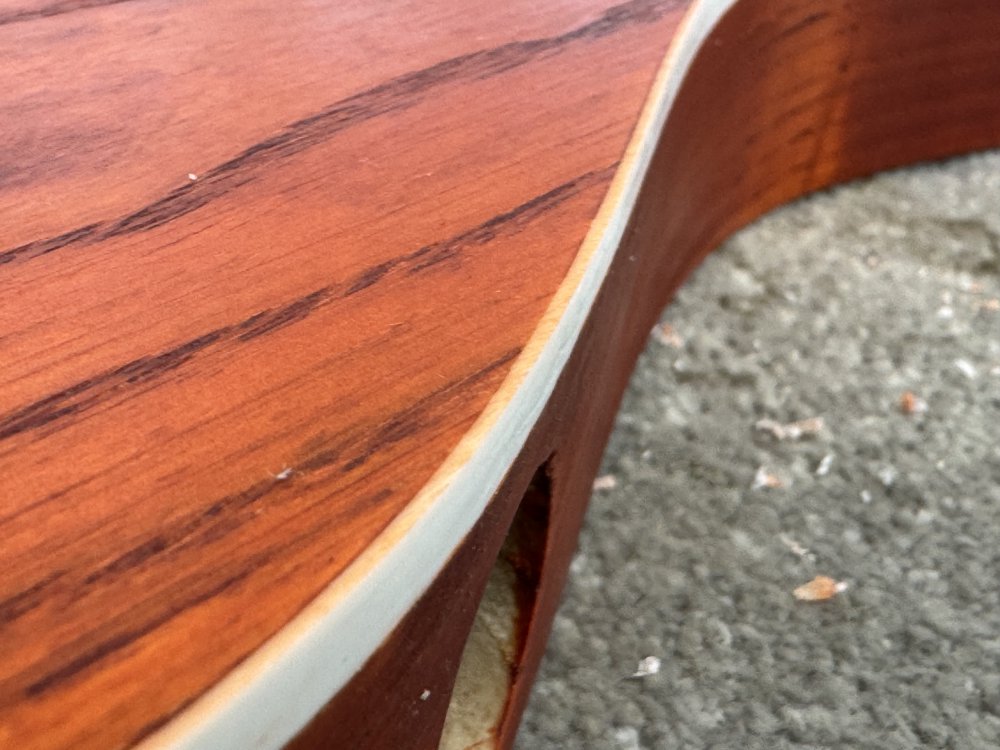 A macro photo of the binding on the edge of a guitar body. The binding as seen from the side of the guitar is about 5mm deep and clean white, but the top face has about 1mm of binding visible, and that still has orange stains on it.