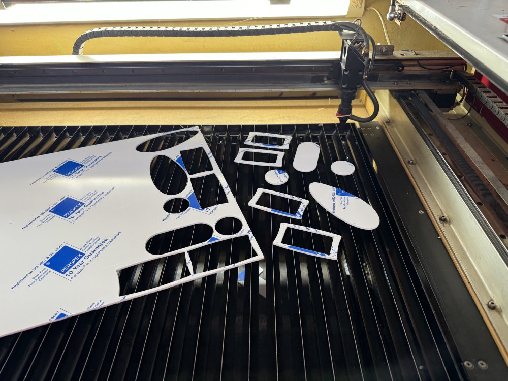 A photo of an industrial looking machine that is a laser cutter. On the bed of the machine is a sheet of white acrylic with various holes in it, and next to that a set of covers and pickup mounts that I cut from the sheet.