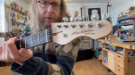 A still from a video showing Michael sat in front of the camera holding a guitar, showing the headstock of it to the camera. On the headstock it says "MWDales" in script and "Delfinen" under that.