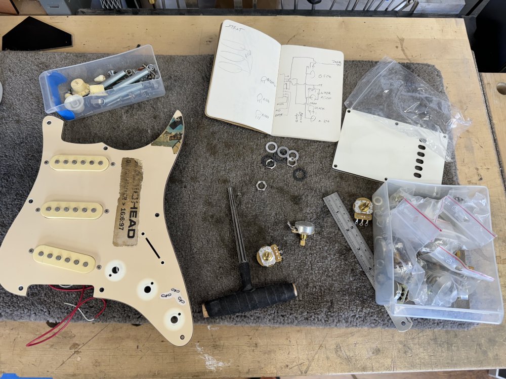 A photo of a pickguard from a stratocaster style guitar on a workbench surrounded by electronics parts and a notebook with a wiring diagram hand draw in it.