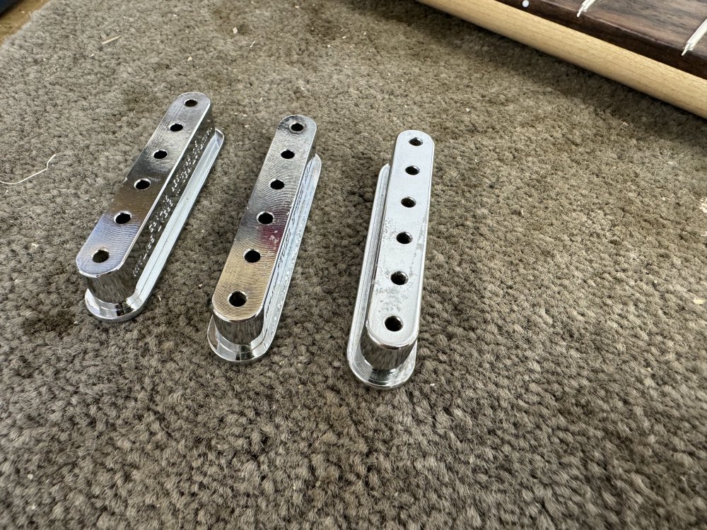 A picture of three metal string ferrule blocks - lozenges about 6 cm long with 6 holes in them for the strings to pass through and a lip on the top edge (currently at the bottom as they're sat upside down on the workbench. If you can look really close you can tell that two of them have been milled on teh bottom whereas the third one is smooth, indicating they were made differently despite otherwise being the same.