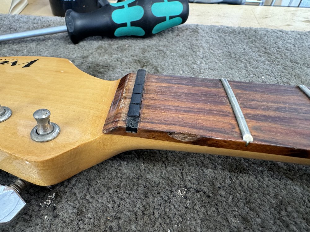 A close-up photo of the nut end of a guitar neck, where there is a black plastic nut with a chip out of it that means the low-E string isn't properly secured.