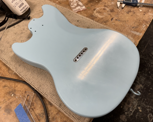 A photo of a blue-painted mustang-style guitar body is sat on the workbench showing a string-ferrule block in place on the rear.