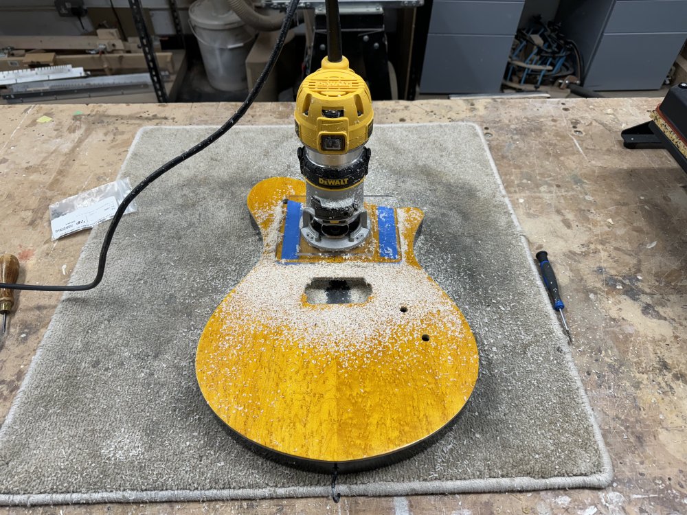 A photo of a yellow and black finished guitar body on a workbench, on top of which is a palm-router and everything is covered in wood-dust from some cutting.