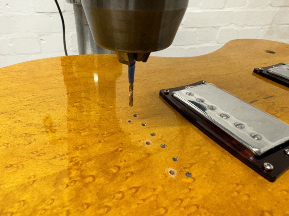 A close-up photo of the yellow topped guitar body on the bed of a pillar-drill, showing a series of holes for where the bridge will go: six holes for the strings through the body and three for the mounting screws. Above of the bridge mounting holes is a drill bit.