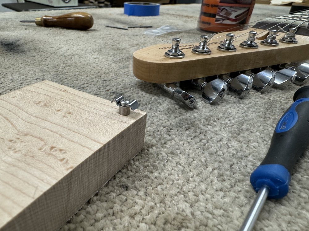 A close up photo showing a string tree screwed into a block of maple wood. Behind it is the neck, awaiting its turn.